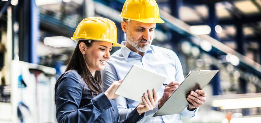 Two workers in yellow hardhats comparing notes on their tablet and clipboard 